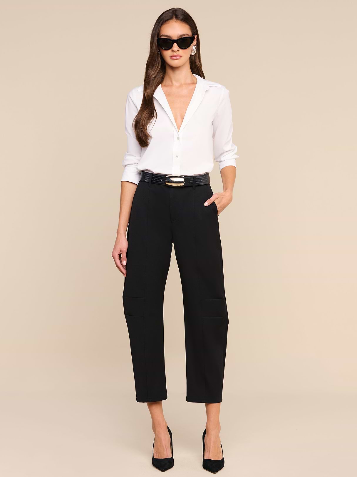 A woman wearing sunglasses, a white button-down shirt, the Colby Barrel-Leg Trouser styled with a black belt, and black heels stands against a beige background with one hand in her pocket.