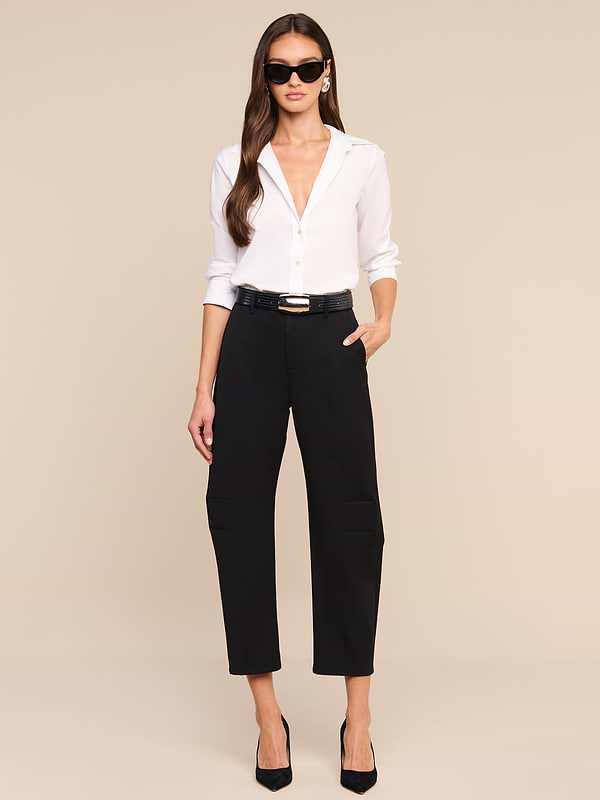 A woman wearing sunglasses, a white button-down shirt, and black heels stands with one hand in her pocket against a plain white background. She completes her effortlessly chic look with the Colby Barrel-Leg Trouser, featuring a modern high-rise barrel-leg silhouette.
