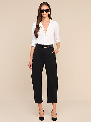 A woman wearing sunglasses, a white button-down shirt, the Colby Barrel-Leg Trouser styled with a black belt, and black heels stands against a beige background with one hand in her pocket.
