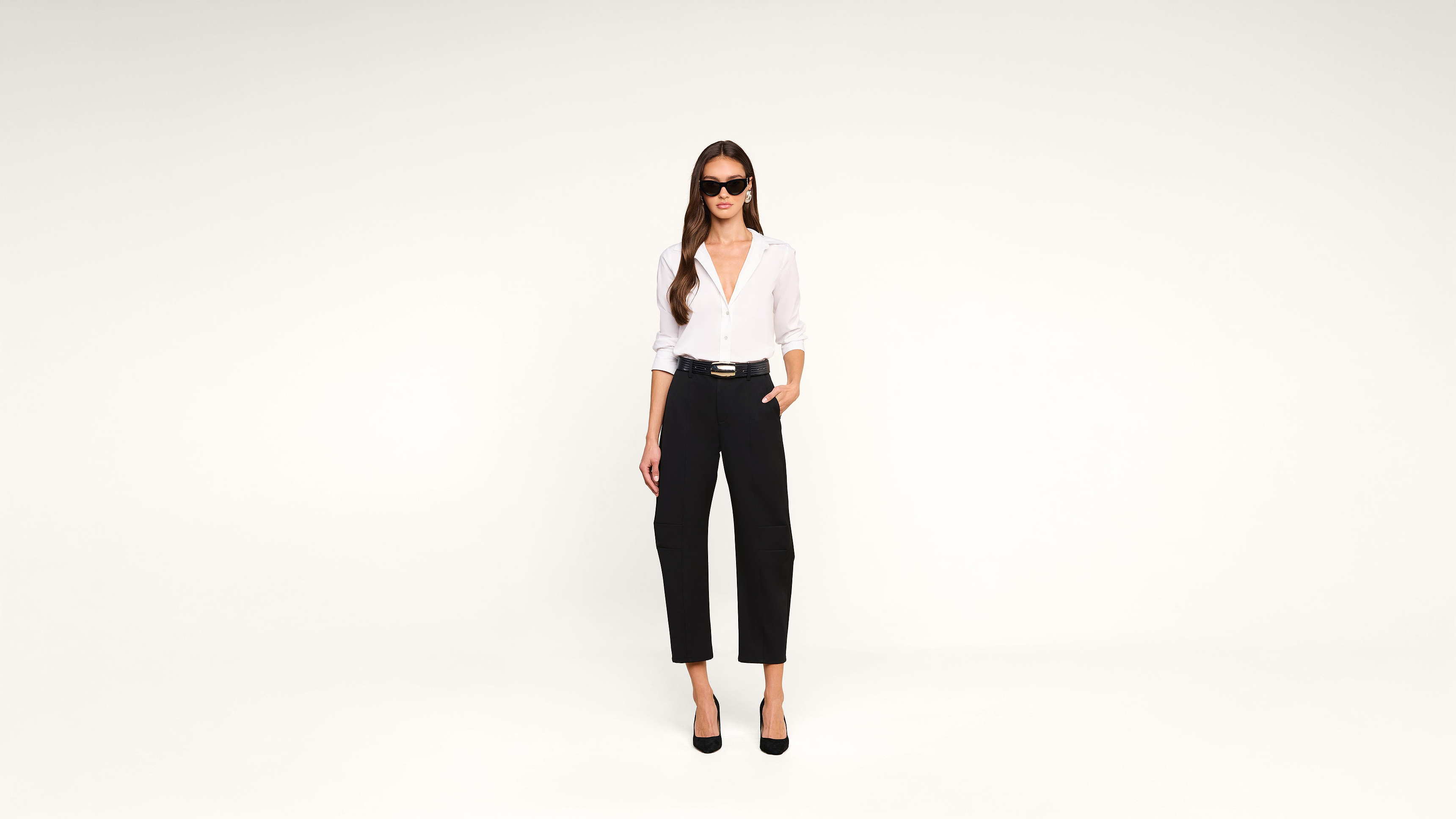 A woman with long hair wears sunglasses, a white blouse, black high-waisted pants, and black heels, standing confidently against a plain white background.
