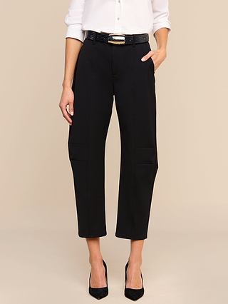 A person stands against a beige background with one hand in their pocket, wearing the Colby Barrel-Leg Trouser in black, styled with a white long-sleeve shirt and black heels.