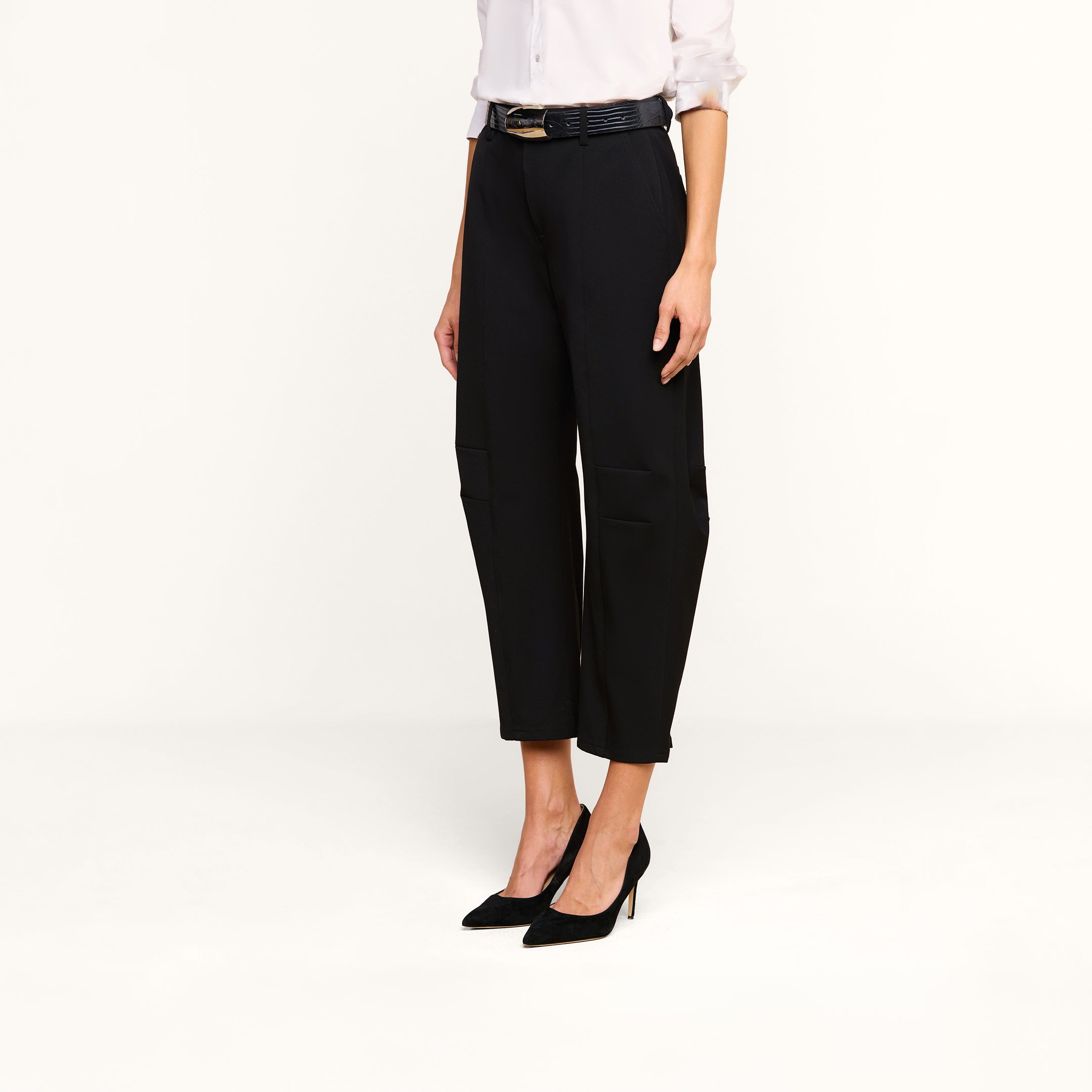 A woman wears high-rise, trouser-style black cropped dress pants—the Colby Barrel-Leg Trouser—paired with a white blouse and belt, complemented by black high heels. She stands against a plain white background, with her face not visible.