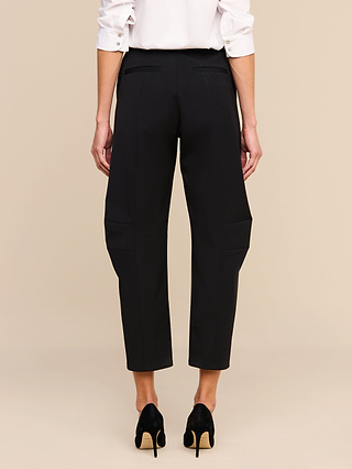 A view from behind of a person wearing a white button-down shirt, the Colby Barrel-Leg Trouser in black with distinctive side-thigh detailing and a subtle barrel-leg silhouette, paired with black high-heeled pumps against a plain beige background.