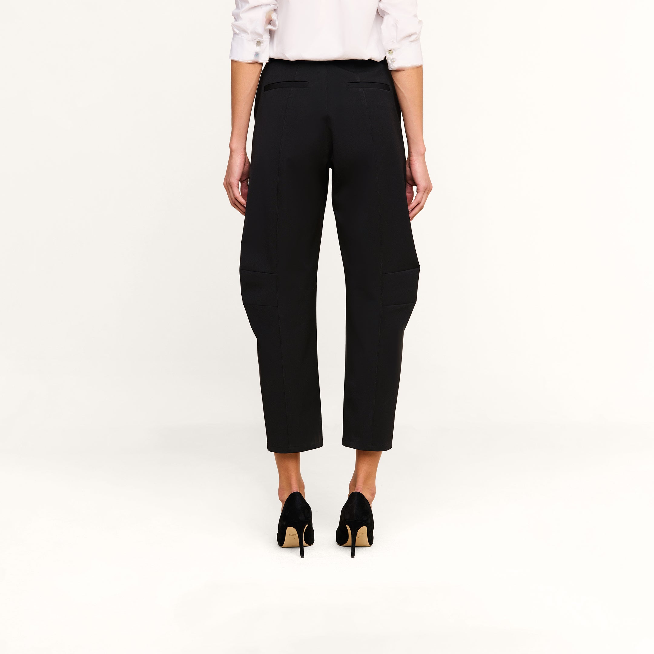 A person stands facing away against a plain white background, wearing the Colby Barrel-Leg Trouser in black, paired with a white long-sleeve shirt with rolled-up sleeves and black high-heeled shoes.