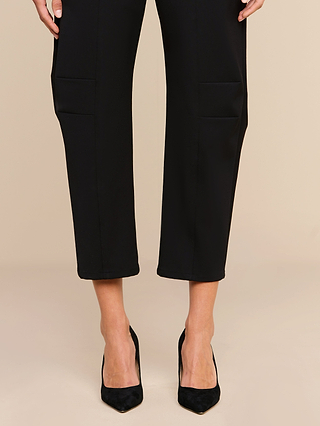 A person wearing the Colby Barrel-Leg Trouser in black, featuring a high-rise fit and side pockets, pairs it with black pointed-toe high heels while standing against a neutral beige background.