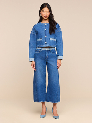 A woman stands facing forward, wearing a cropped denim jacket and the Bronte Cropped Wide-Leg Jean, which features a high waistband and a cropped wide-leg silhouette. She pairs the look with blue heels against a plain beige background.