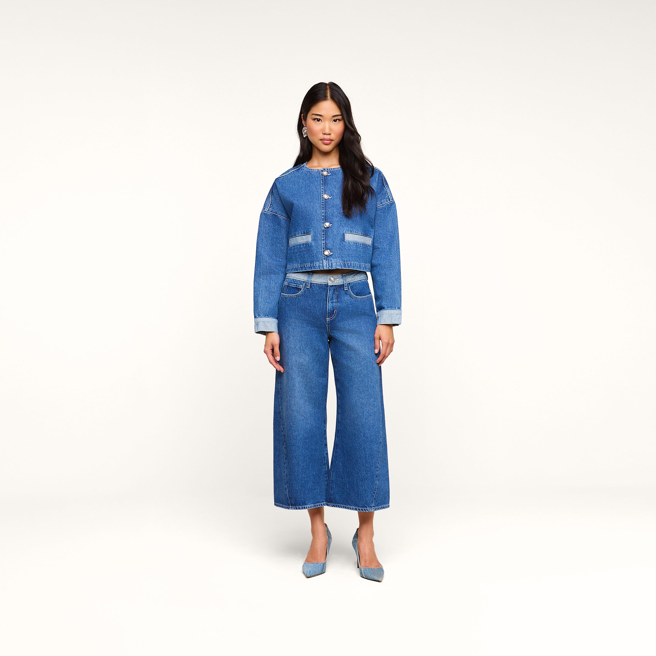 Eine Frau steht mit dem Blick nach vorne und trägt eine kurze Jeansjacke sowie die „Bronte Cropped Wide-Leg Jean“, die sich durch einen hohen Bund und eine kurze Silhouette mit weitem Bein auszeichnet. Vor einem schlichten beigen Hintergrund kombiniert sie den Look mit blauen Pumps.