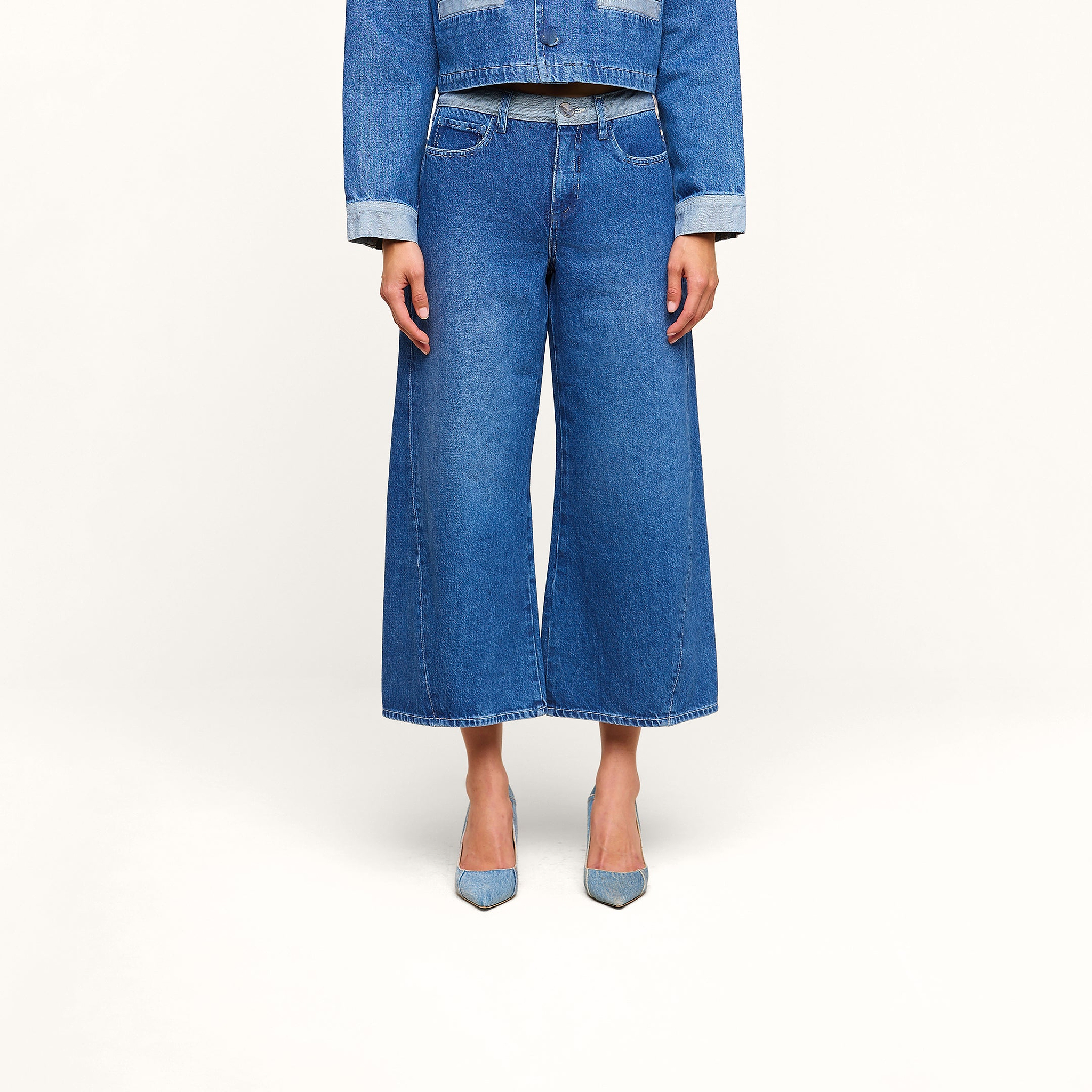 Eine Person trägt eine blaue Jeansjacke mit hochgekrempelten Ärmeln, dazu eine „Bronte Cropped Wide-Leg Jeans“ mit hohem Bund. Zu sehen ist nur der Oberkörper von den Schultern bis zu den Füßen; das Outfit wird durch hellblaue Pumps mit spitzer Zehenpartie abgerundet.