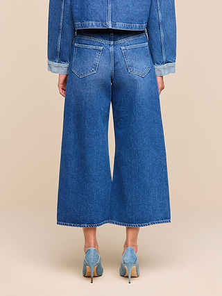 Model wearing the Bronte Cropped Wide-Leg Jean with a high waistband, paired with a denim jacket and light blue high-heeled shoes, shown from the back against a plain beige background.