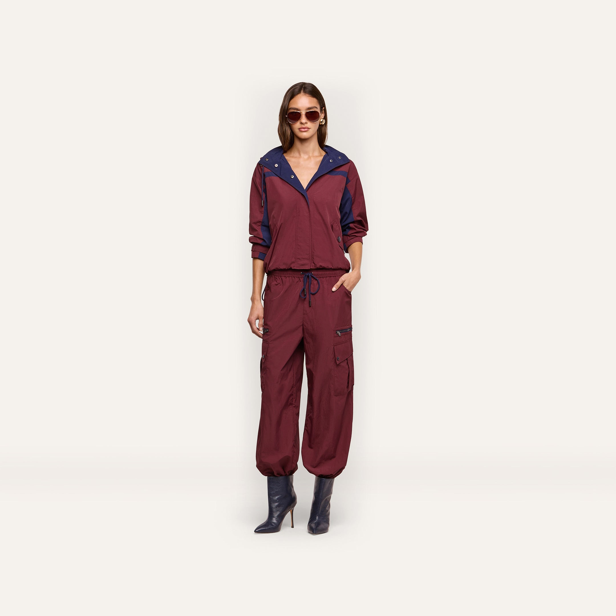 A woman wears a maroon water-repellant hooded jacket and Monrose Nylon Jogger cargo pants with dark blue accents, paired with dark heeled boots and sunglasses.