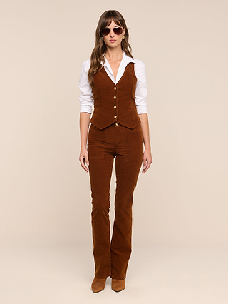 A woman wearing a white shirt, brown high rise stretch corduroy vest, matching Saffia Corduroy Pant, brown shoes, and sunglasses stands facing forward against a beige background.
