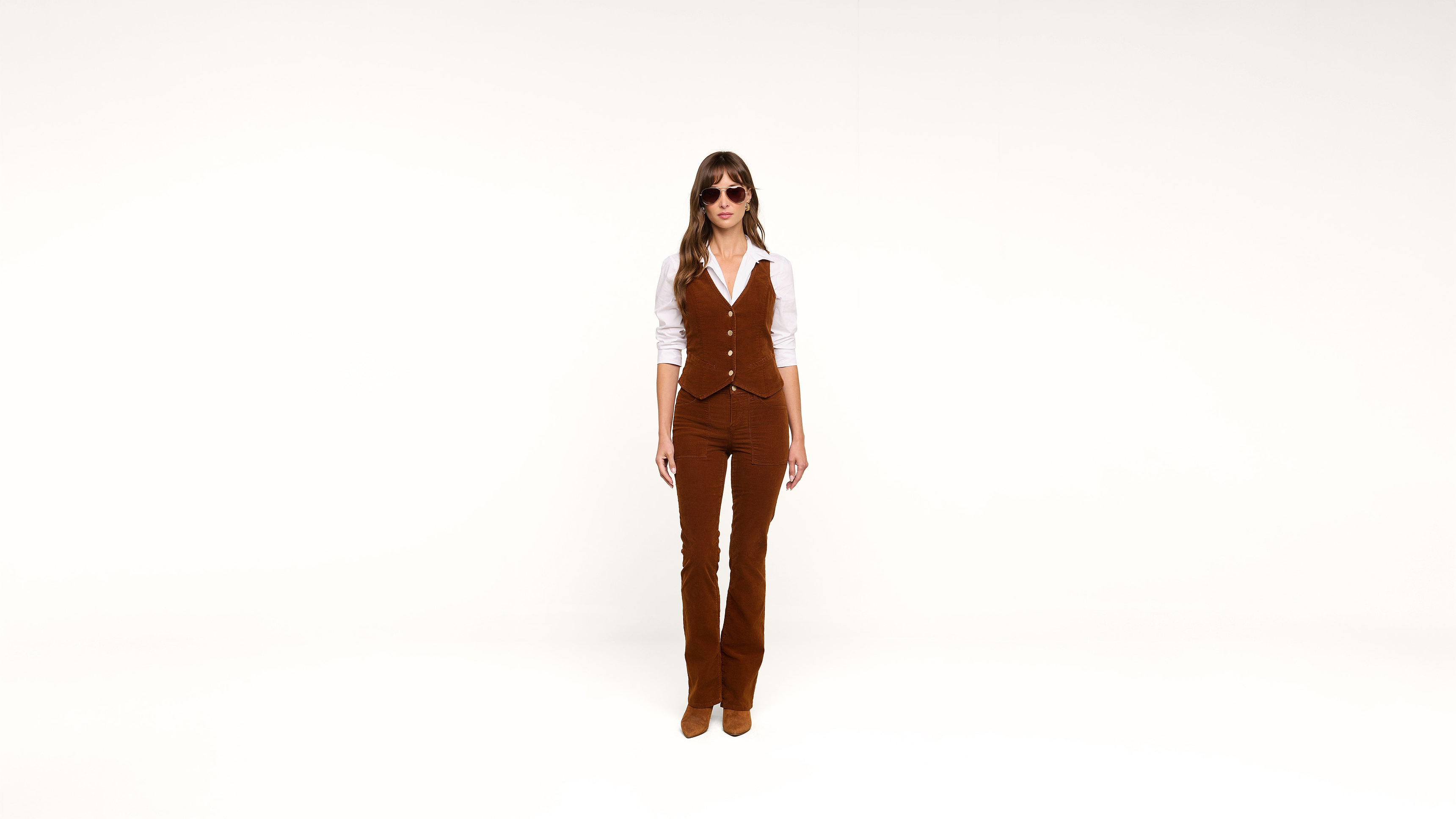 A person stands against a plain white background, wearing brown flared pants, a matching vest, a white shirt, and sunglasses.