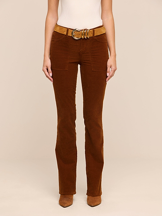 A woman wearing the Saffia Corduroy Pant—high-rise, slim-straight stretch corduroys with front patch pockets—paired with a tan suede belt featuring a large gold buckle and matching brown suede boots, stands against a neutral background.