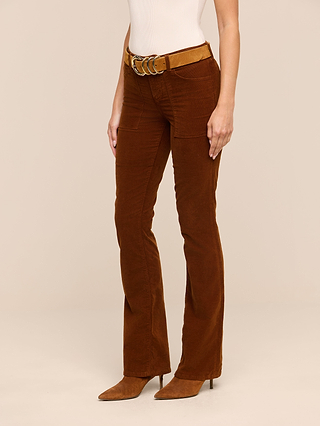 A person wearing the Saffia Corduroy Pant, featuring a high rise, brown stretch corduroy fabric and front pockets, styled with a tan belt, white sleeveless top, and brown pointed-toe shoes, stands against a neutral background.