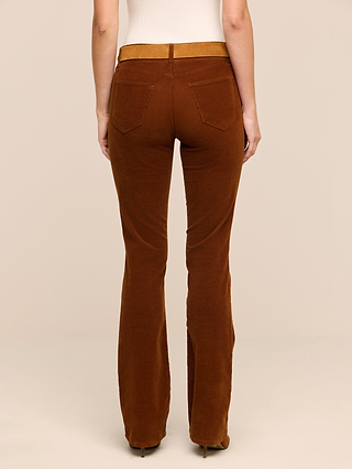 Someone is shown from the back, wearing the Saffia Corduroy Pant—high-waisted brown stretch corduroy pants—standing against a plain light beige background with only their lower torso and legs visible.