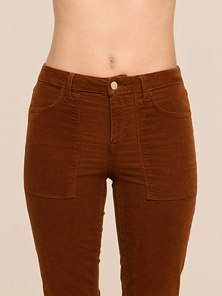 A person wearing the Saffia Corduroy Pant—a high-waisted, brown stretch corduroy style with large front pockets—shown from the waist to mid-thigh against a plain background.