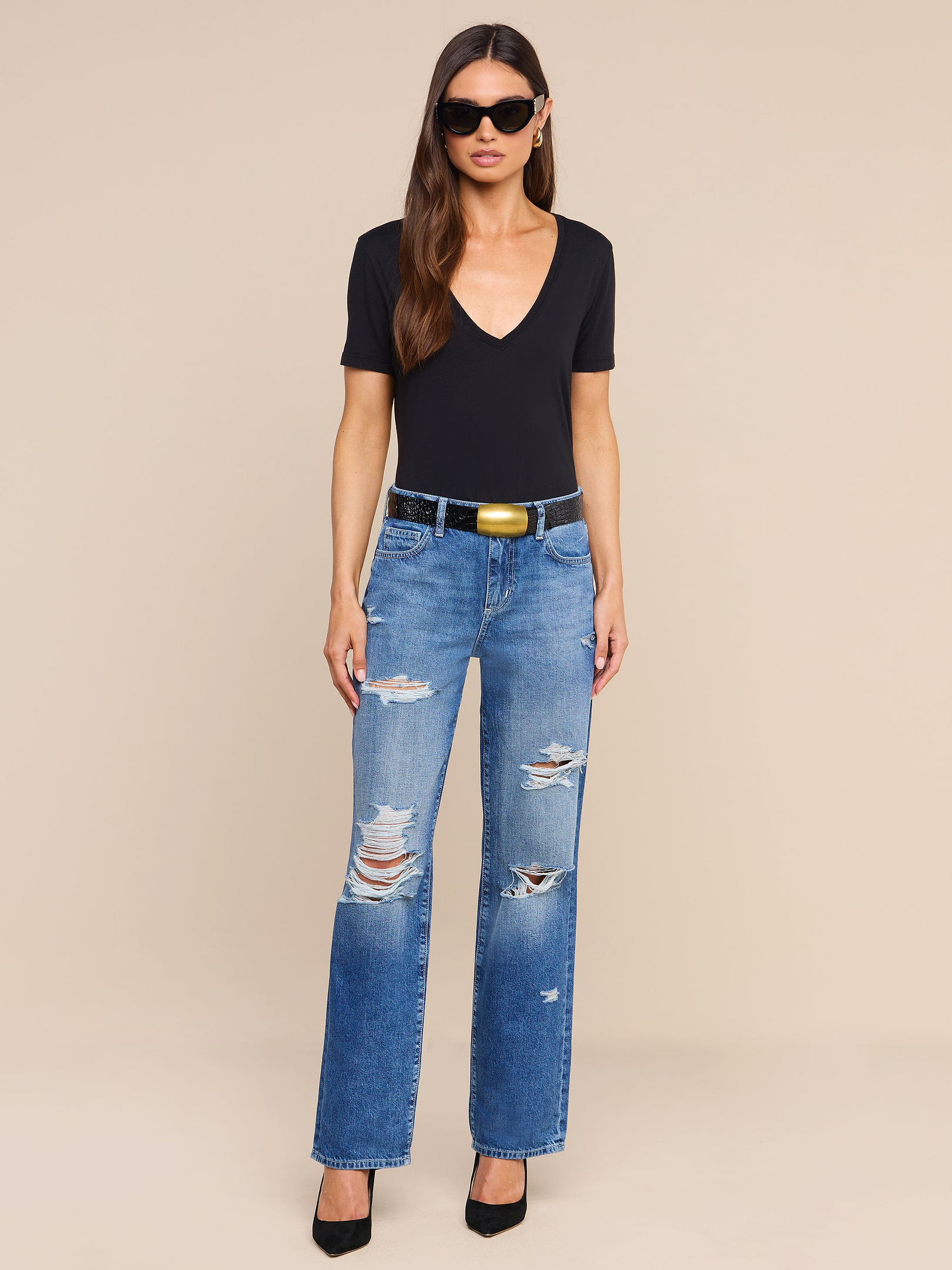 A woman stands against a plain white background, wearing sunglasses, a black short-sleeve V-neck top, and the Zelda Slouchy Straight Jean in destructed blue denim. She completes her look with a black belt featuring a gold buckle and black heels.