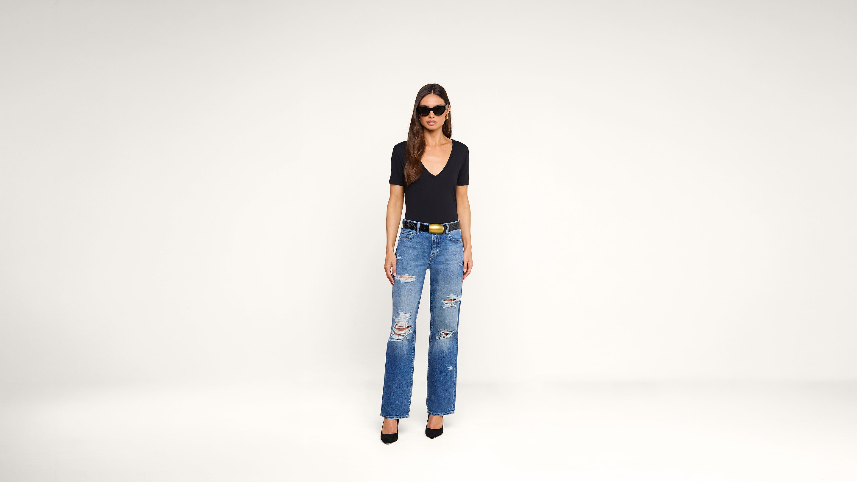 Woman wearing sunglasses, a black V-neck top, ripped blue jeans, and black heels stands against a plain white background.