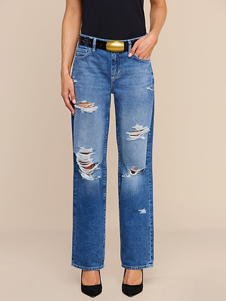 A person stands against a plain beige background wearing the Zelda Slouchy Straight Jean in a medium indigo wash, styled with a black belt featuring a gold buckle, a black top, and black heels. The effortless cool edge of the look comes from the slouchy-straight silhouette of the Zelda jeans.