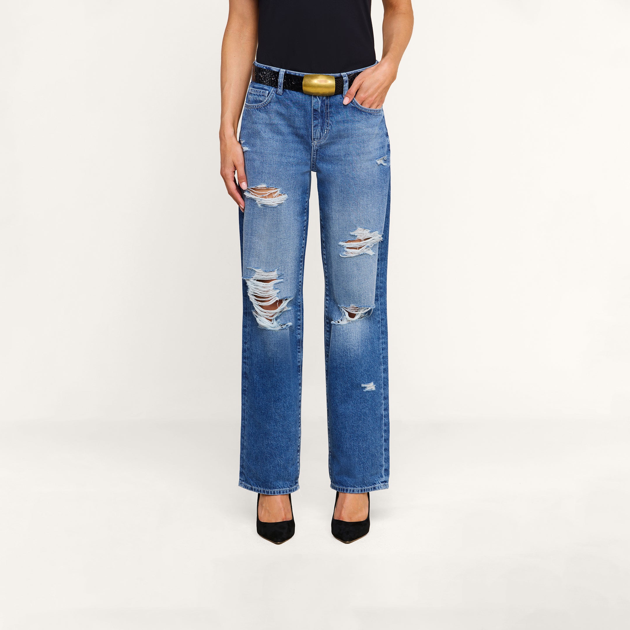 A person wearing the Zelda Slouchy Straight Jean in a destructed, ripped blue denim style, paired with a black top, a black belt with a gold buckle, and black heels stands against a plain white background.