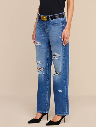 Wearing the Zelda Slouchy Straight Jean in a medium indigo wash with rips on both legs, styled with a black belt, black top, and black pointed-toe heels, the person stands against a plain beige background.