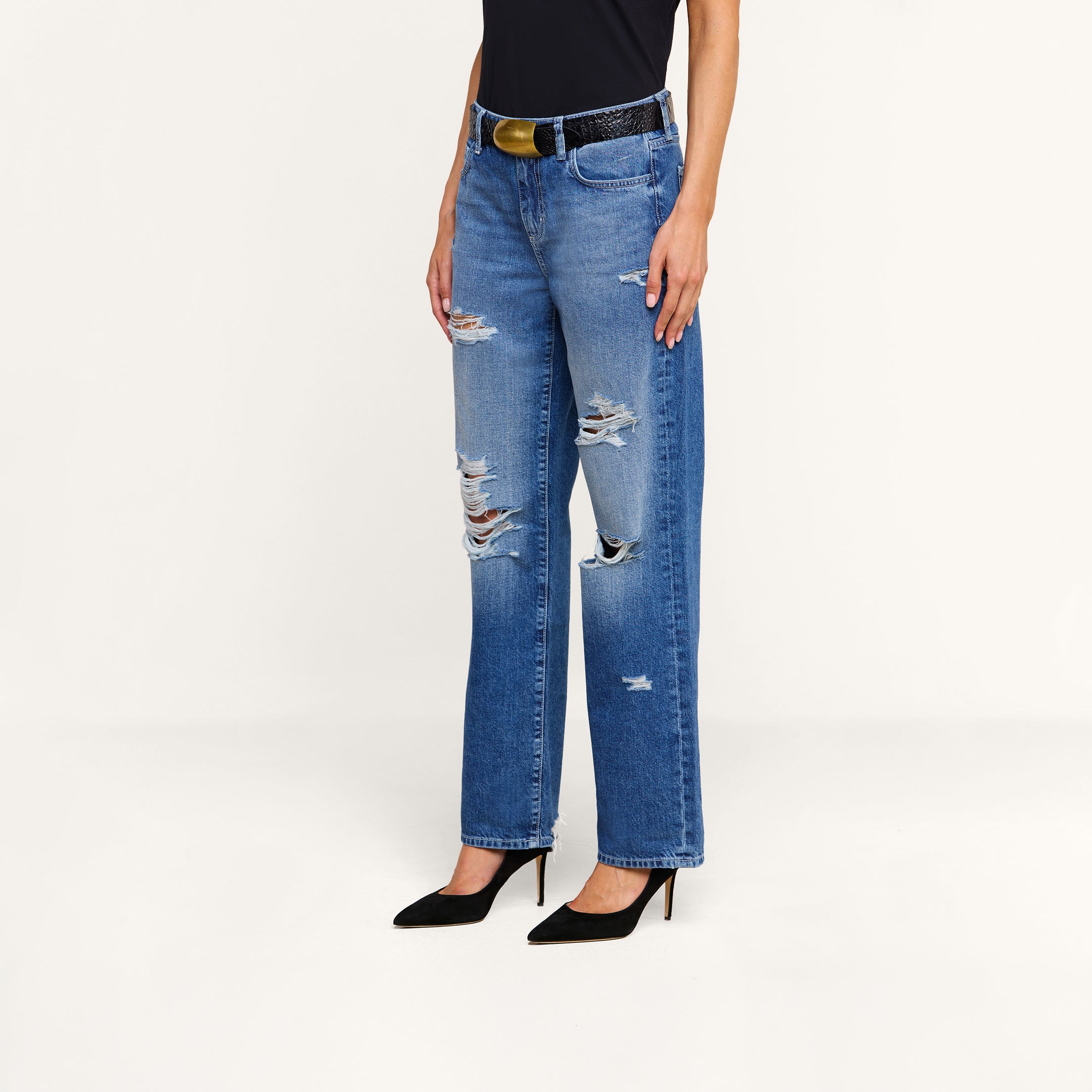 A person wearing a black top, the Zelda Slouchy Straight Jean in destructed denim, a black belt, and black high heels stands against a plain white background.