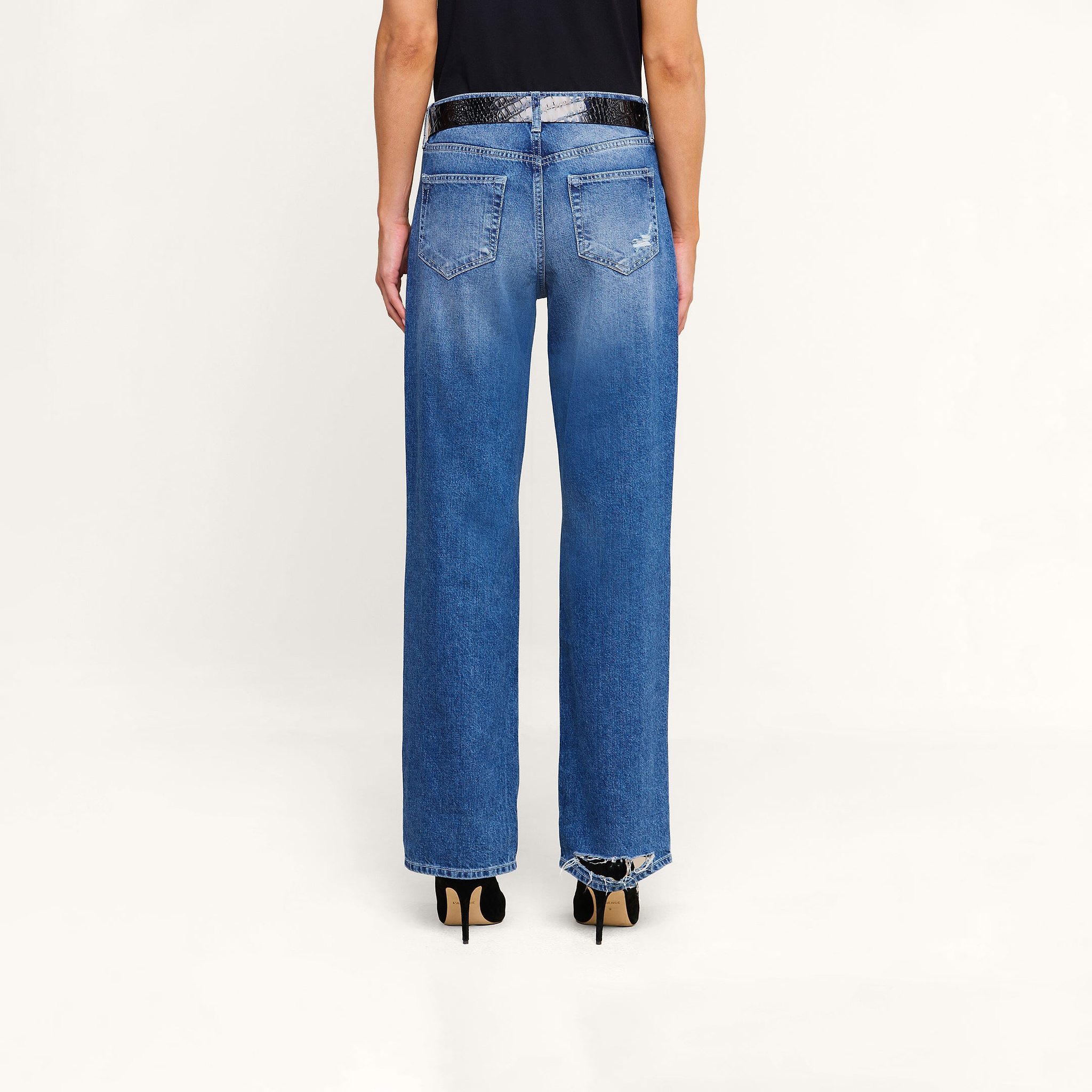 A person stands against a white background, wearing the Zelda Slouchy Straight Jean with a frayed hem, paired with a black shirt and black high-heeled shoes. The image displays a back view from waist to feet.