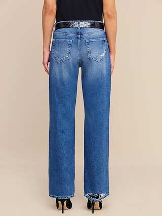 Wearing the Zelda Slouchy Straight Jean in a medium indigo wash, this person showcases its relaxed fit, slouchy-straight silhouette, and visible stitching. The jeans have subtle distressing near the back pocket and are styled with a black textured belt and black high heels. The look is set against a plain beige background.