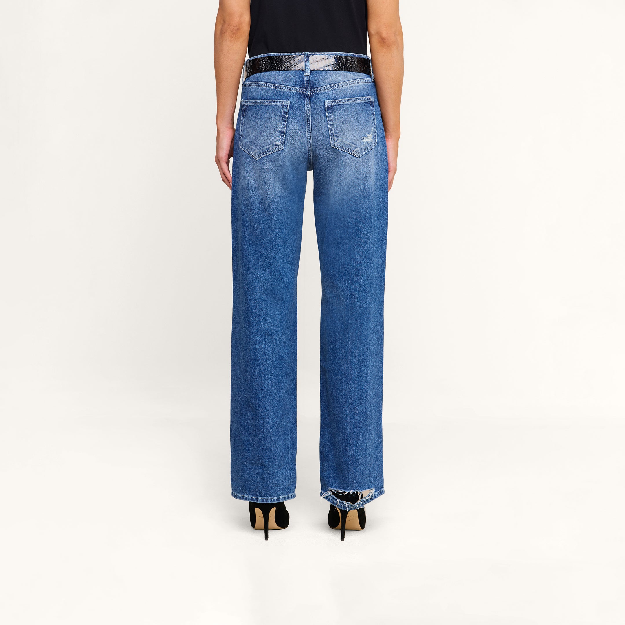 A person stands against a white background, wearing the Zelda Slouchy Straight Jean with a frayed hem, paired with a black shirt and black high-heeled shoes. The image displays a back view from waist to feet.