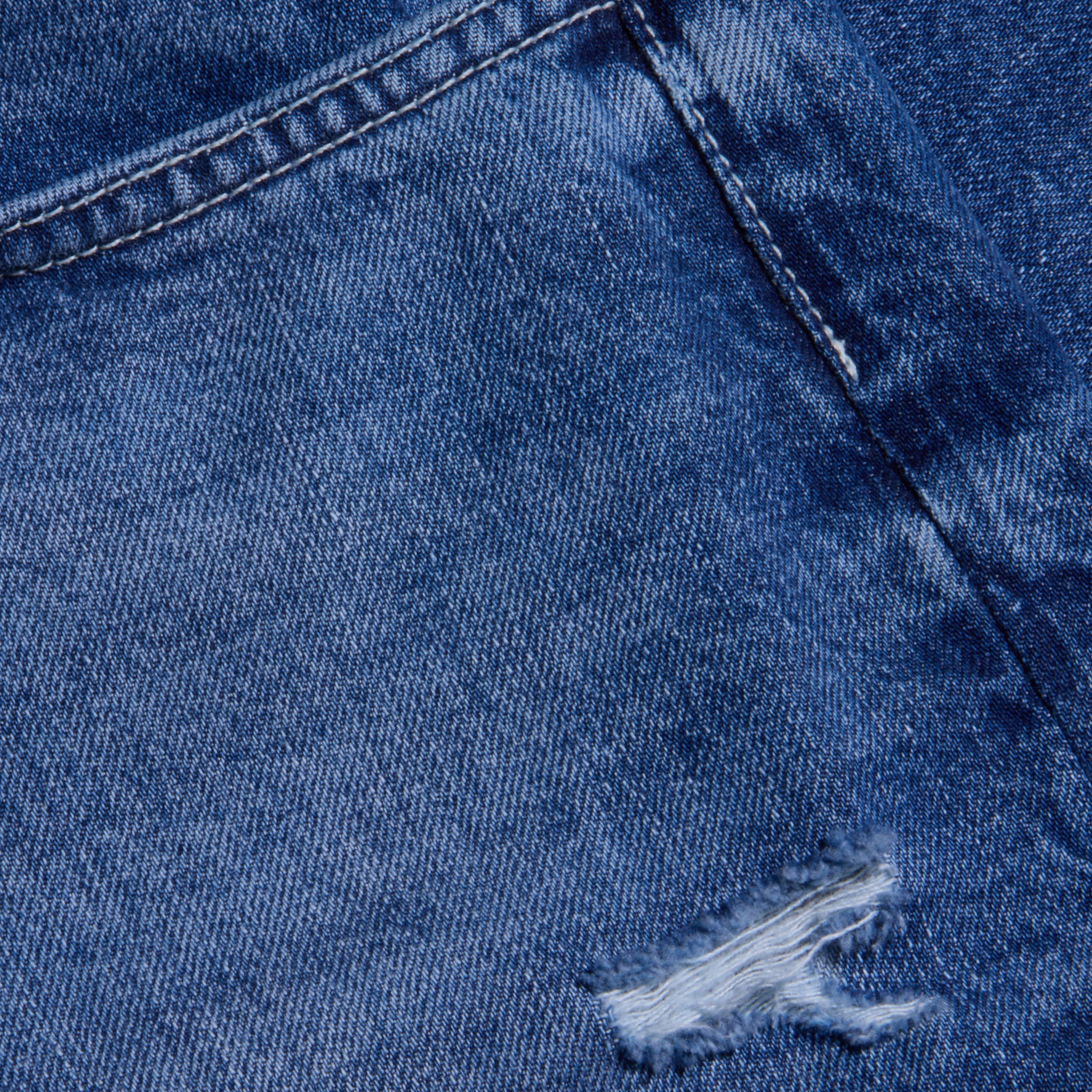 Close-up view of blue destructed denim fabric showing a seam and a small ripped, frayed hole, highlighting the texture and worn appearance of the Zelda Slouchy Straight Jean.