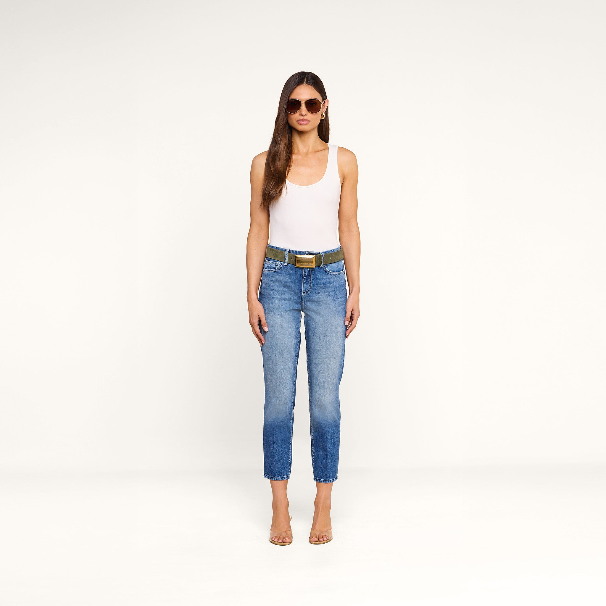 A woman stands against a plain white background, facing forward with her arms relaxed at her sides, wearing sunglasses, a white tank top, clear heels, and the Avena Slim-Leg Jean with a belt.