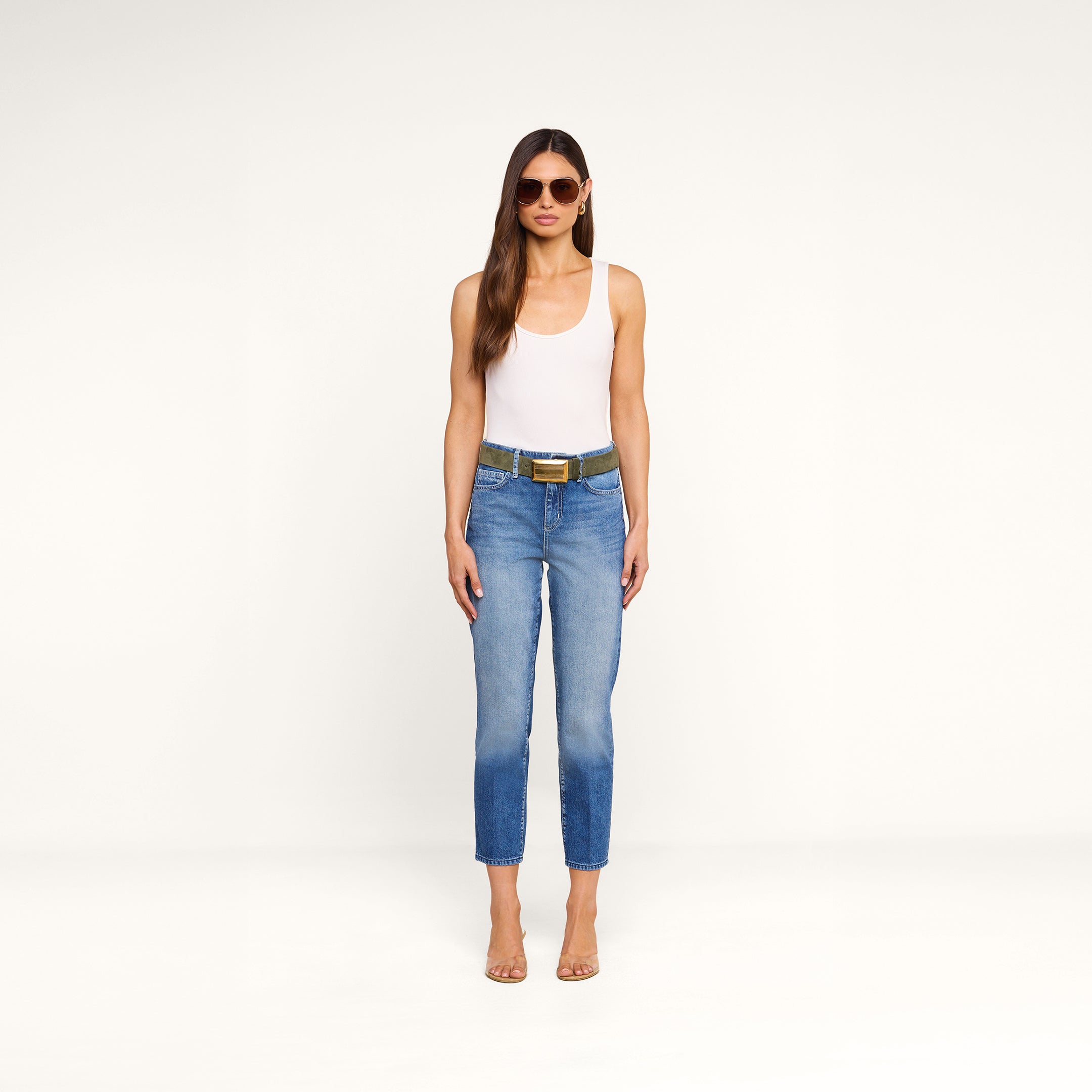 A woman stands against a plain white background, facing forward with her arms relaxed at her sides, wearing sunglasses, a white tank top, clear heels, and the Avena Slim-Leg Jean with a belt.