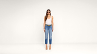 A woman stands against a plain white background, wearing sunglasses, a white tank top, blue jeans with a belt, and clear-heeled sandals. She has long brown hair and her arms are relaxed at her sides.