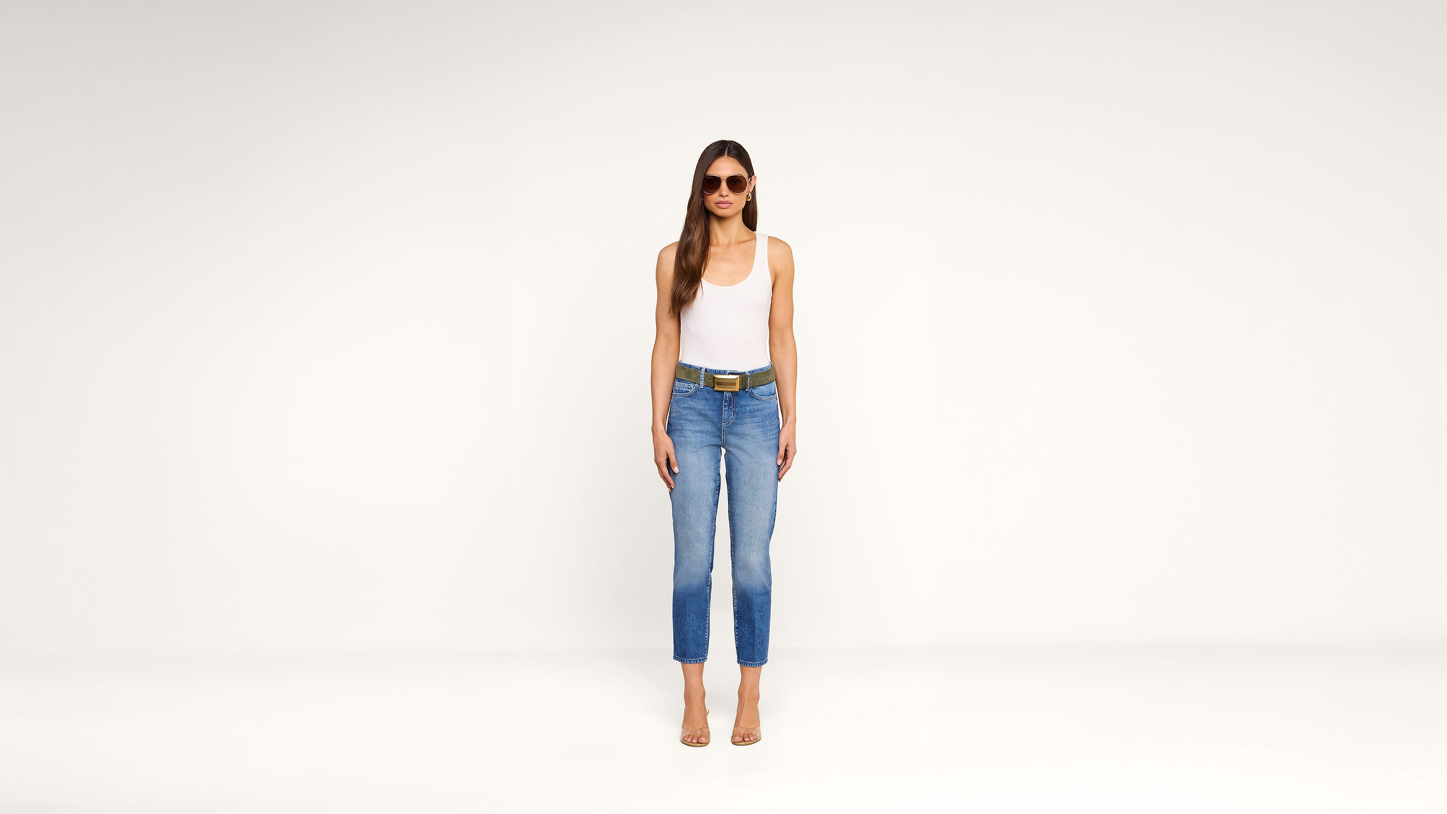 A woman stands against a plain white background, wearing sunglasses, a white tank top, blue jeans with a belt, and clear-heeled sandals. She has long brown hair and her arms are relaxed at her sides.
