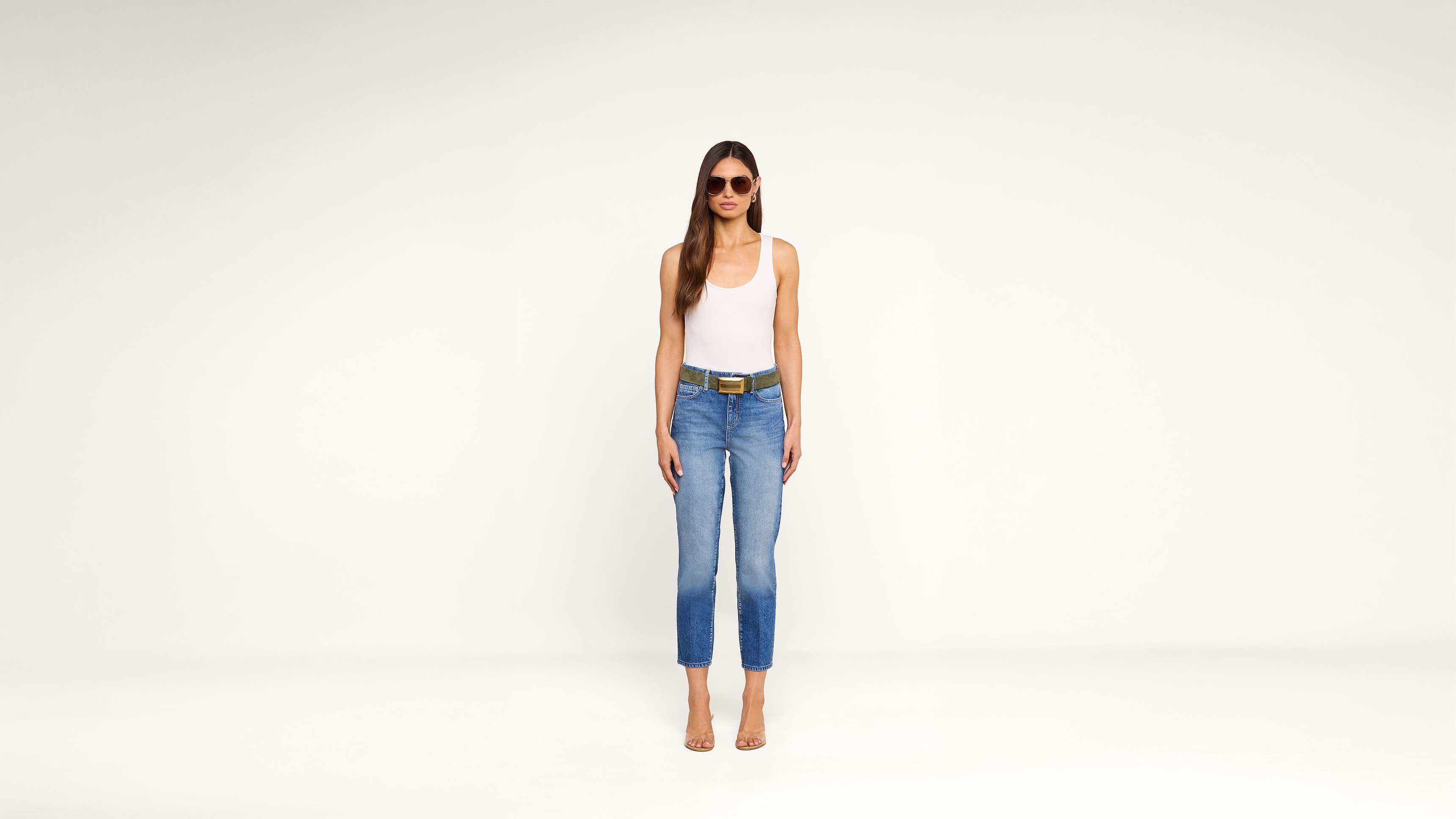 A woman stands against a plain white background, wearing sunglasses, a white tank top, blue jeans with a belt, and clear-heeled sandals. She has long brown hair and her arms are relaxed at her sides.