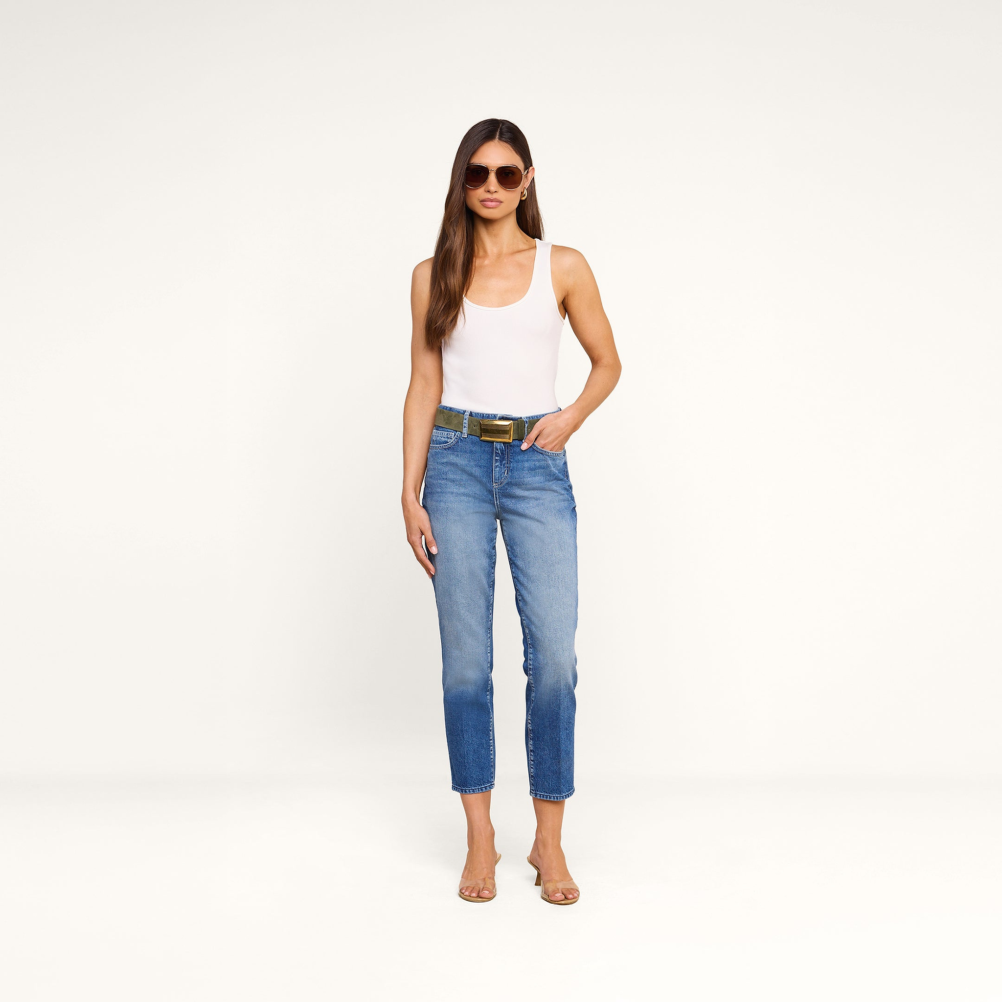 A woman wearing sunglasses, a white tank top, the Avena Slim-Leg Jean, and heeled sandals stands against a plain white background with her hands in her pockets.