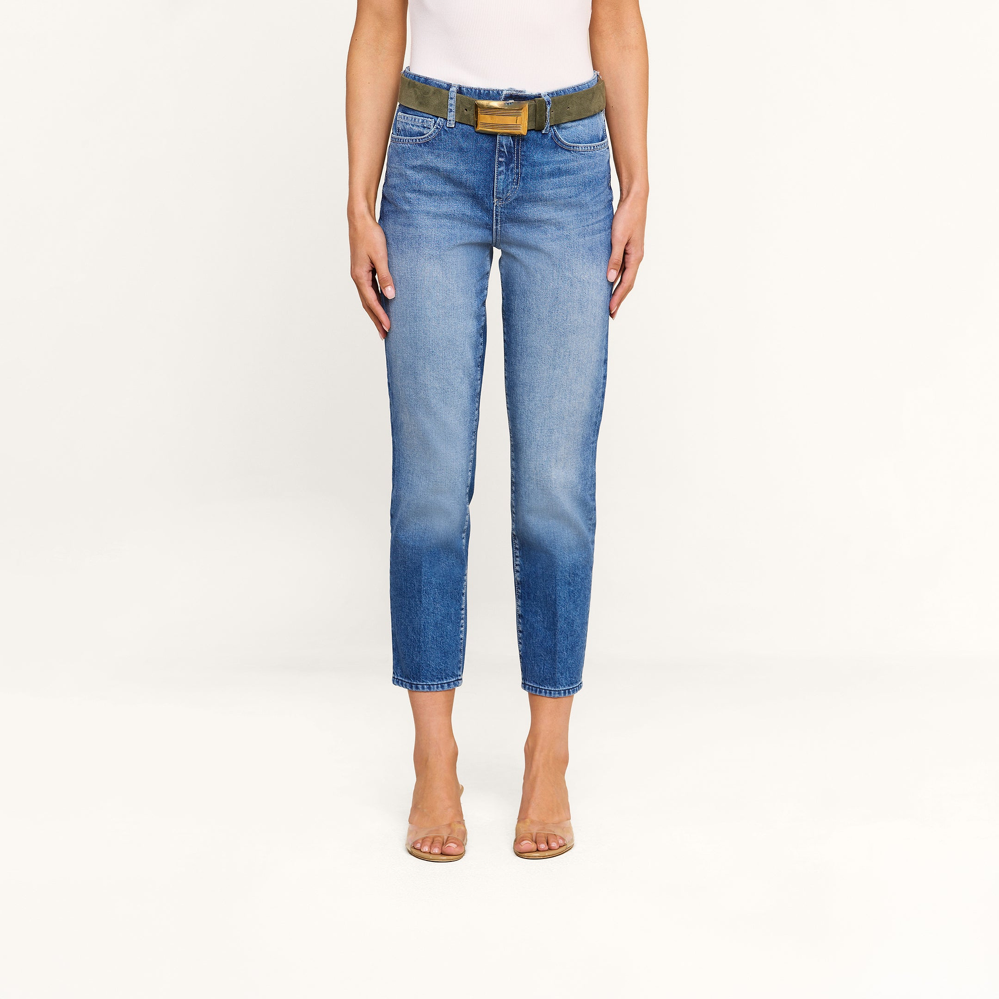 A person wearing a white top, the Avena Slim-Leg Jean in blue with a green belt featuring a brown buckle, and clear high-heeled shoes stands against a plain white background.