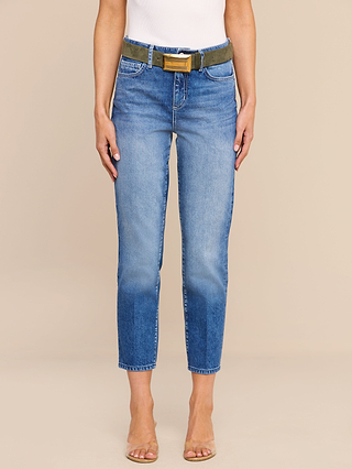 A person wearing blue Avena Slim-Leg Jeans, a white top, a green belt with a gold buckle, and clear heeled sandals stands against a neutral background.