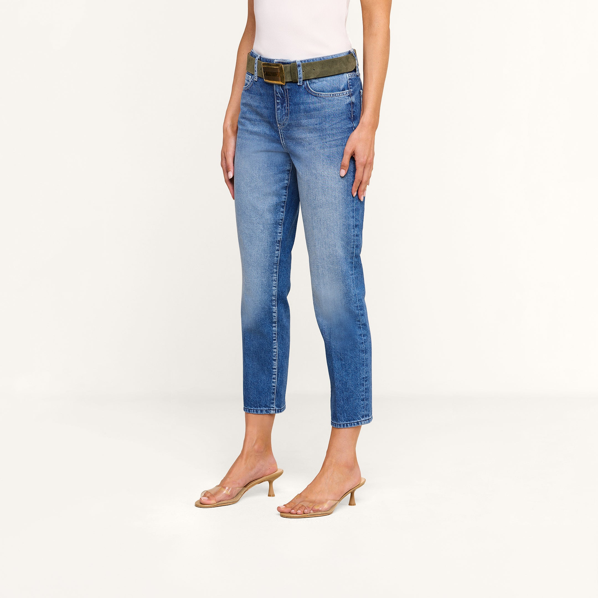 A person wearing the Avena Slim-Leg Jean in denim blue, a dark belt, a white sleeveless top, and clear heeled sandals stands against a plain, light background.