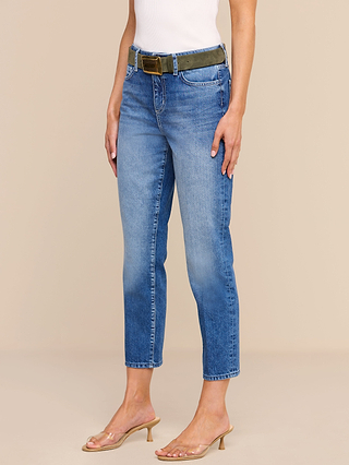 A person wearing the Avena Slim-Leg Jean in blue, a white sleeveless top, an olive green belt, and clear heeled sandals stands against a beige background. Their head is not visible in the image.