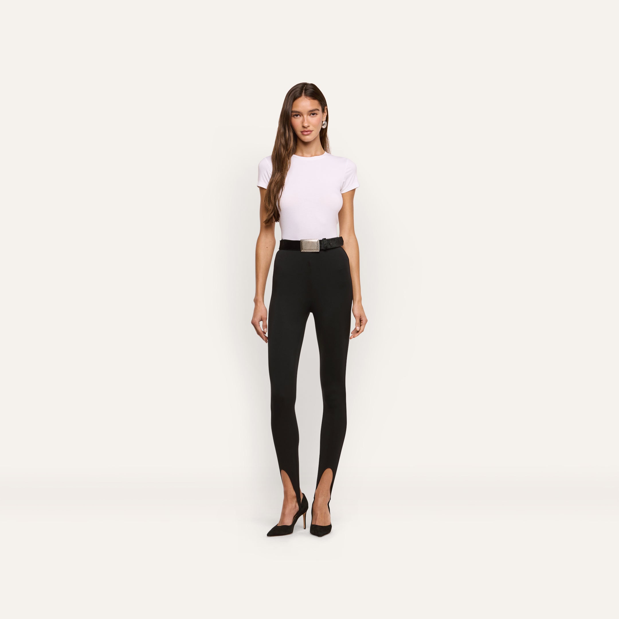 A woman wearing a white t-shirt, Enid Stirrup Legging in black scuba knit, a black belt, and black heels.