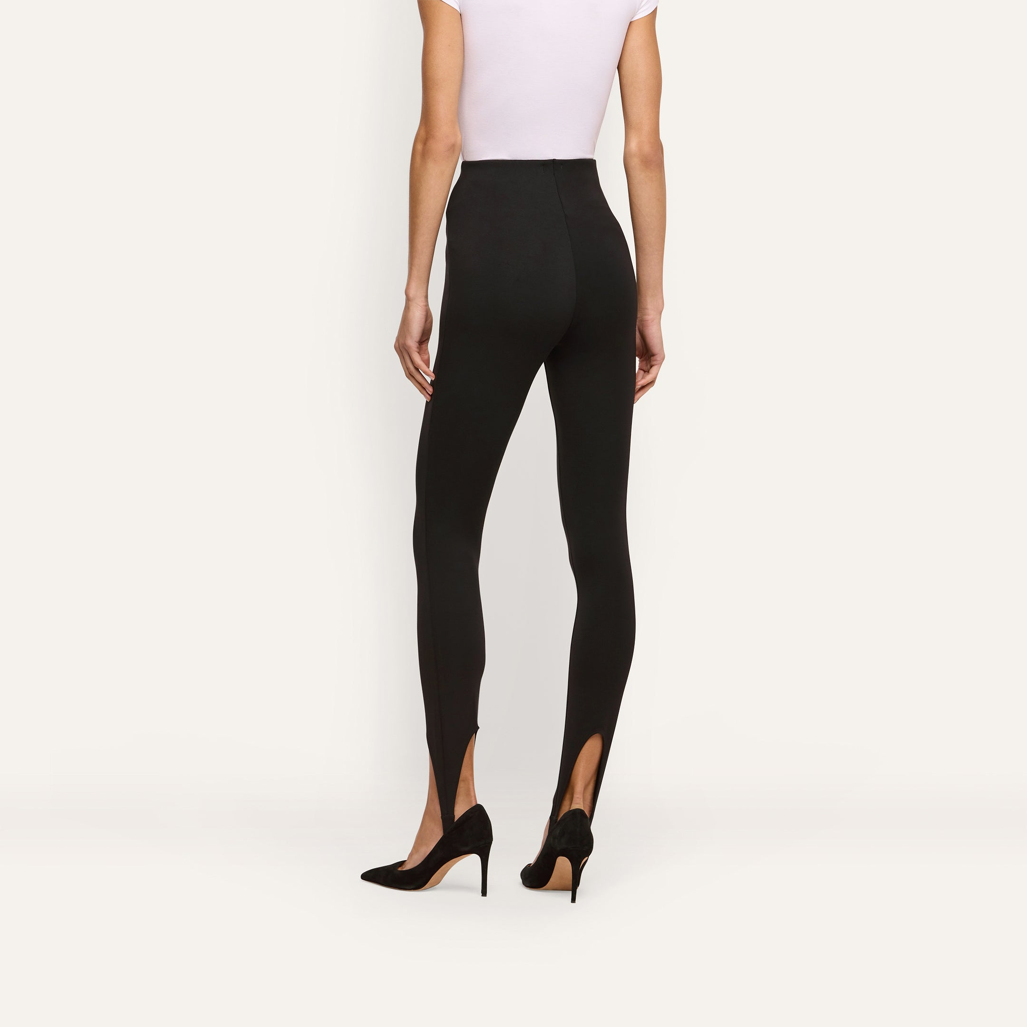 Back view of a person wearing black high-rise Enid Stirrup Legging scuba knit leggings with black pointed-toe heels.