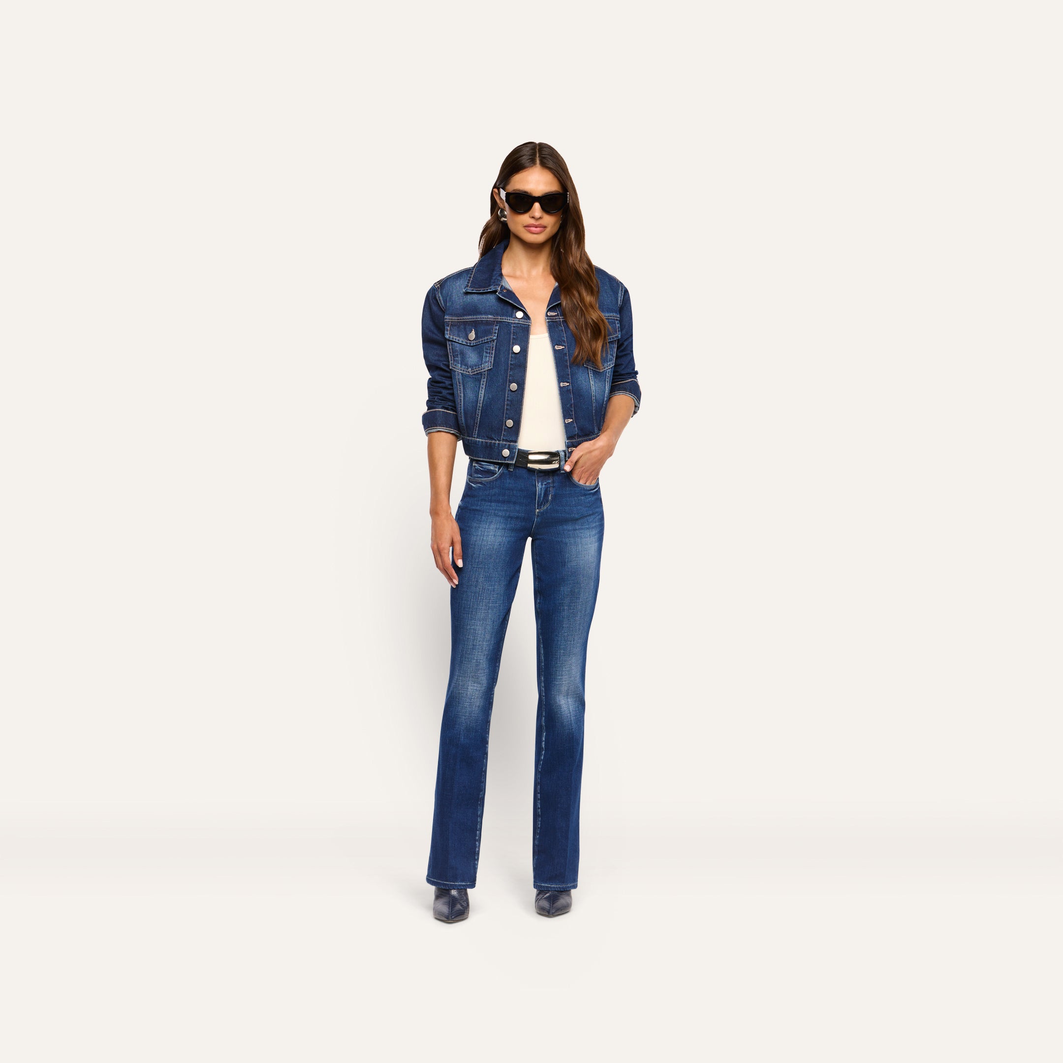 A woman wearing a dark blue denim jacket over a white top, and Kezia Bootcut Jean low-rise stretch jeans with a black belt.