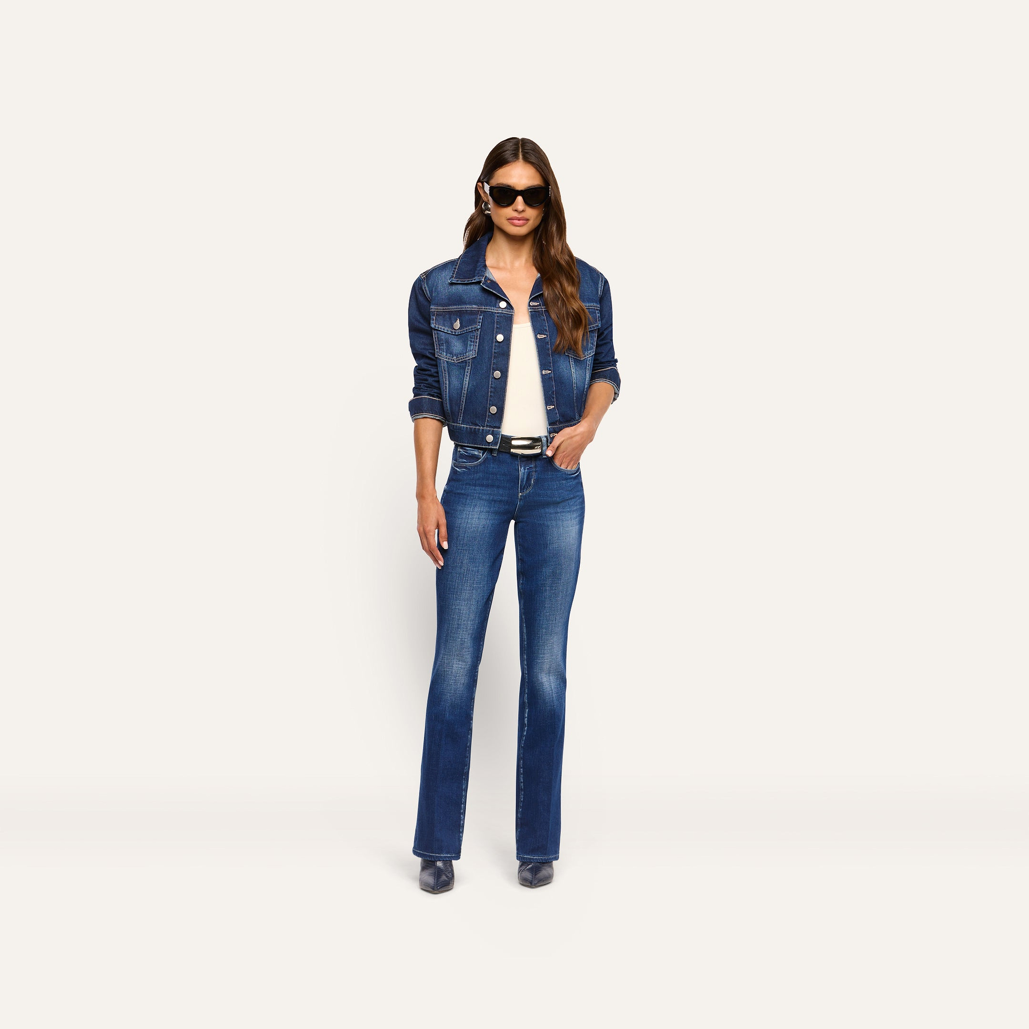 A woman wearing a dark blue denim jacket over a white top, and Kezia Bootcut Jean low-rise stretch jeans with a black belt.