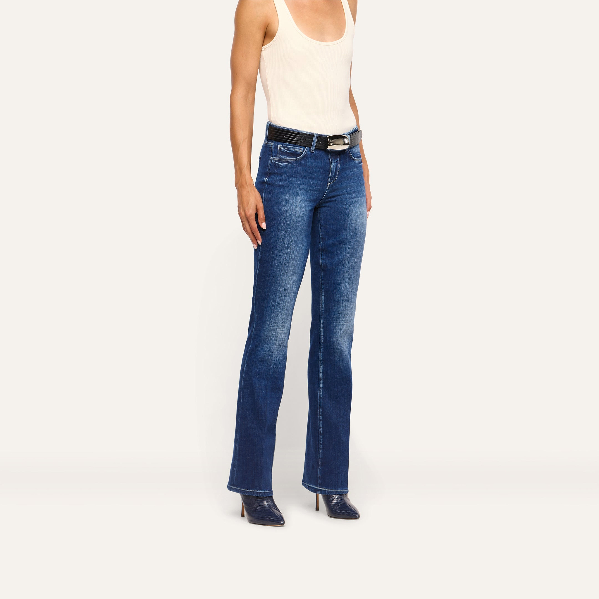 A person wears a beige tank top and Kezia Bootcut Jean low-rise jeans with a black belt and dark pointed-toe heels.