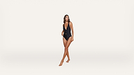 A woman wearing a black one-piece swimsuit stands barefoot against a plain white background, looking off to the side with one hand resting at her waist.