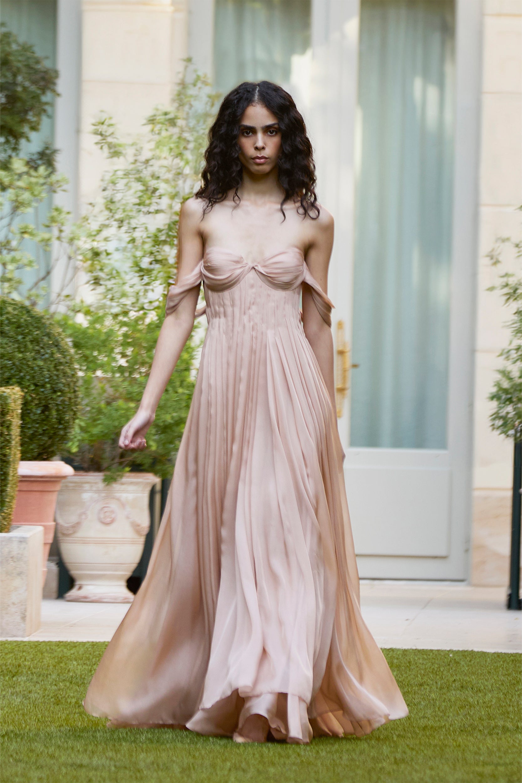A person with long dark hair wears a flowing, off-shoulder, light pink gown while walking outdoors on grass, surrounded by greenery and vibrant potted plants.