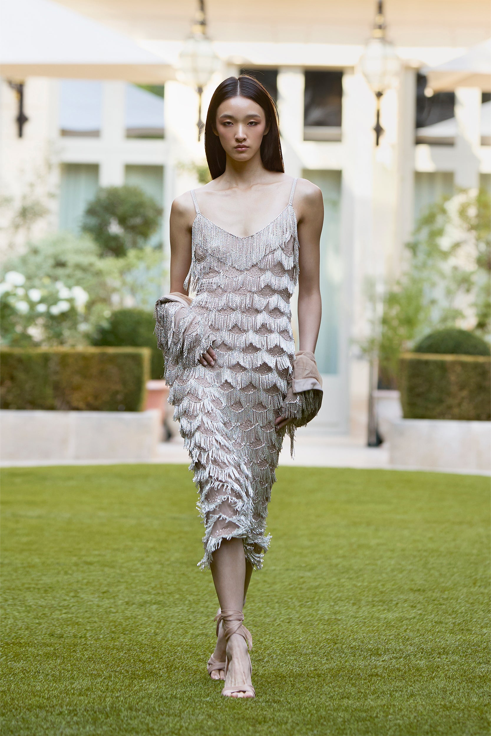 A model in a silver, fringed, sleeveless dress and strappy sandals walks outside on green grass, framed by manicured hedges and modern buildings in the background.