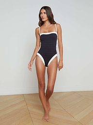 A woman stands barefoot against a plain light background wearing the Tory Bandeau One-Piece Swimsuit in navy with white trim, featuring thin shoulder straps and a high leg cut.