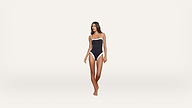 A woman stands barefoot against a plain white background, wearing a navy blue one-piece swimsuit with white trim.